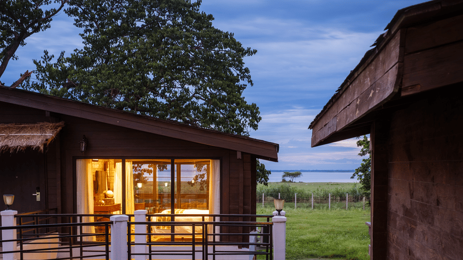 A close-up of a wooden cabin at Rhino & River Wildlife Retreat & Spa, Pobitora, featuring large glass windows glowing with warm light against the evening landscape.
