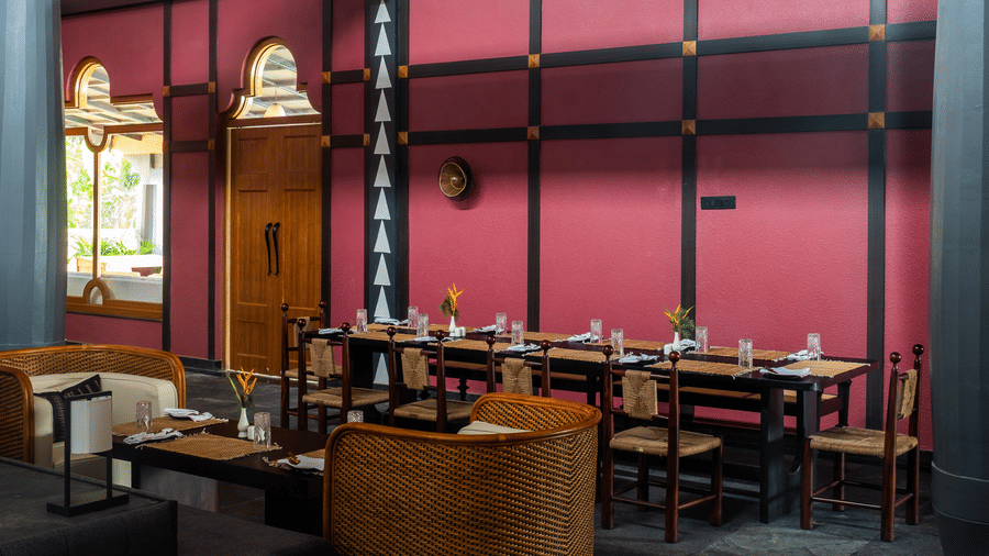 A wide view of the dining area at Rhino & River Wildlife Retreat & Spa, Pobitora, featuring long wooden tables, wicker chairs, and deep red walls with black decorative framing.