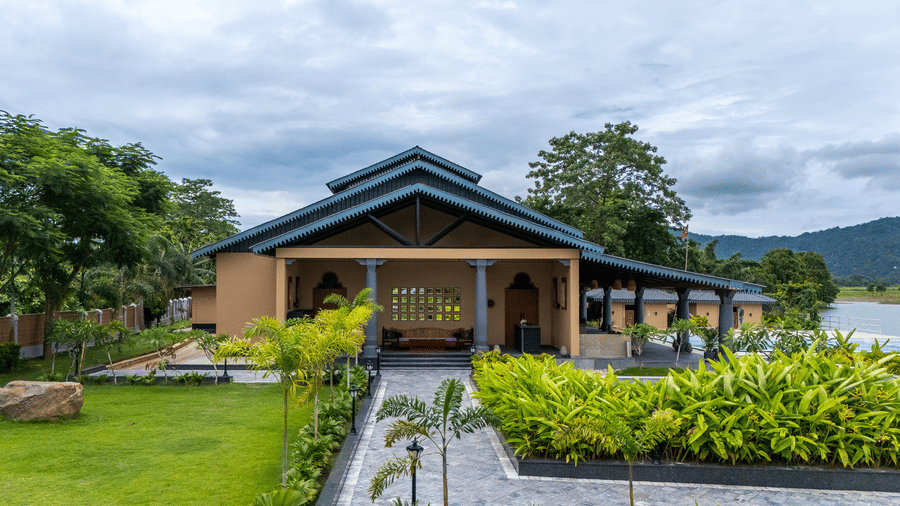 The restaurant building of Rhino & River Wildlife Retreat & Spa, Pobitora, featuring a large gabled roof, manicured green lawn, and lush shrubbery.