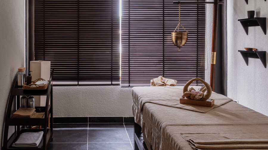 A serene spa treatment room with a massage table, wooden blinds, and hanging brass oil lamps at Rhino & River Wildlife Retreat & Spa, Pobitora.