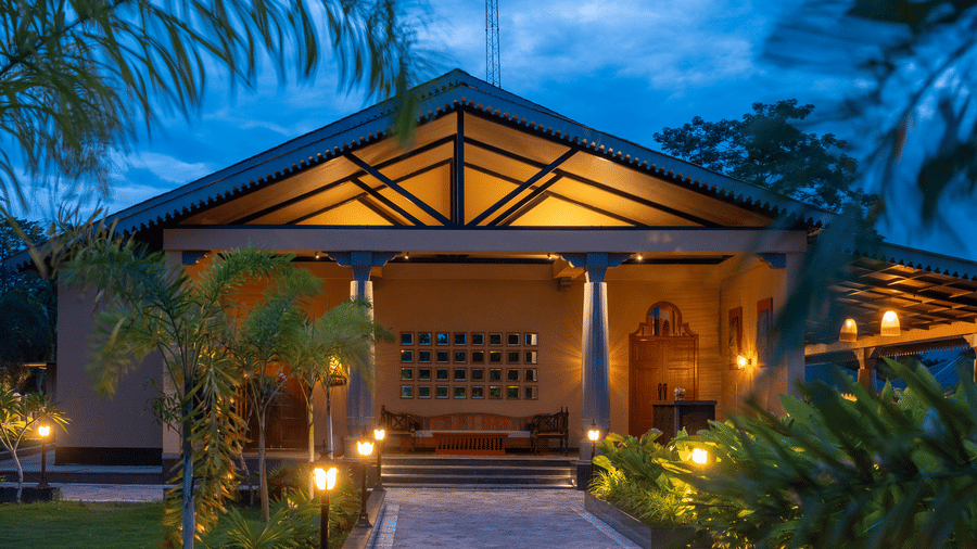The illuminated entrance of a villa at Rhino & River Wildlife Retreat & Spa, Pobitora, at dusk, showing warm interior lighting, a gabled roof, and landscaped palm trees.