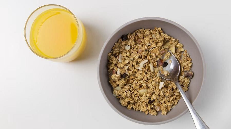 a glass of orange juice and a bowl of granola with a spoon, on a white surface.