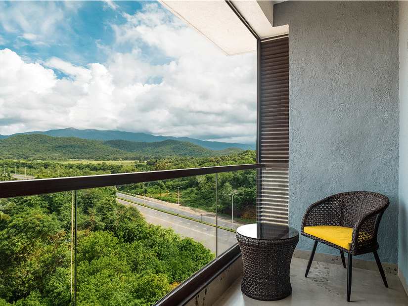  A chair and coffee table on the balcony of one of the rooms offer a view of lush trees and mountains under a cloudy sky at Cloud9 Sarovar Premiere, Palolem Goa