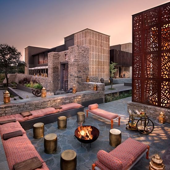 An image of an open courtyard where a few stools, chairs, a bed, and a rack with liquid items are placed and in the middle there is fire burning - Aramness