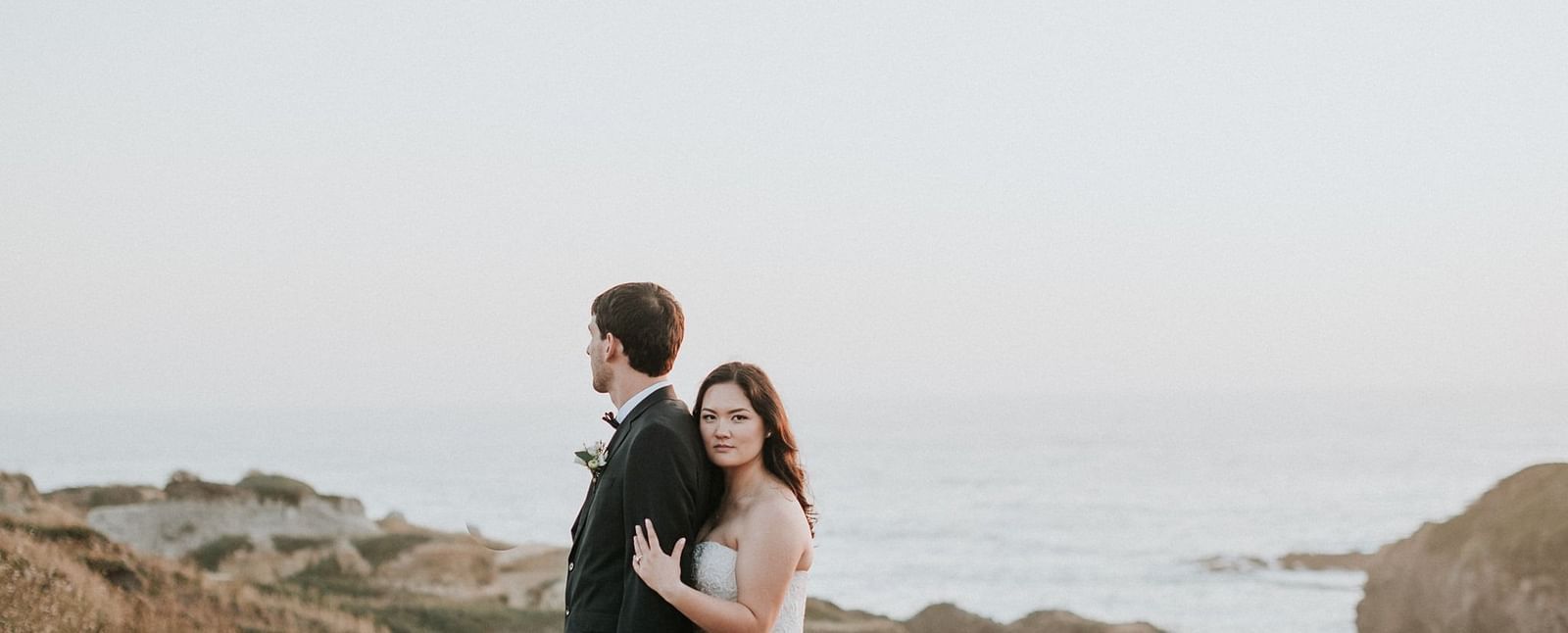 Bride and groom embracing each other on a grassy hill by the ocean