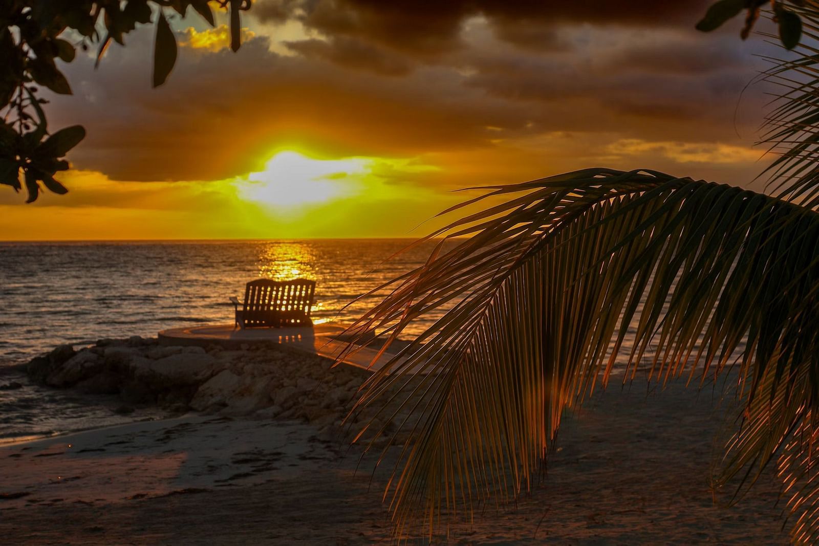 A sunset view over the ocean, with a silhouette of a structure on the horizon and foliage in the foreground.