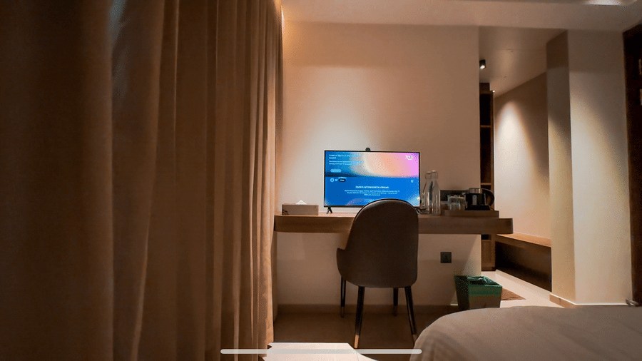 A panoramic shot from the bed of the Deluxe Room featuring a chair and a table with a TV in front of the bed, with blackout curtains covering the windows at F Hotels, Bengaluru.
