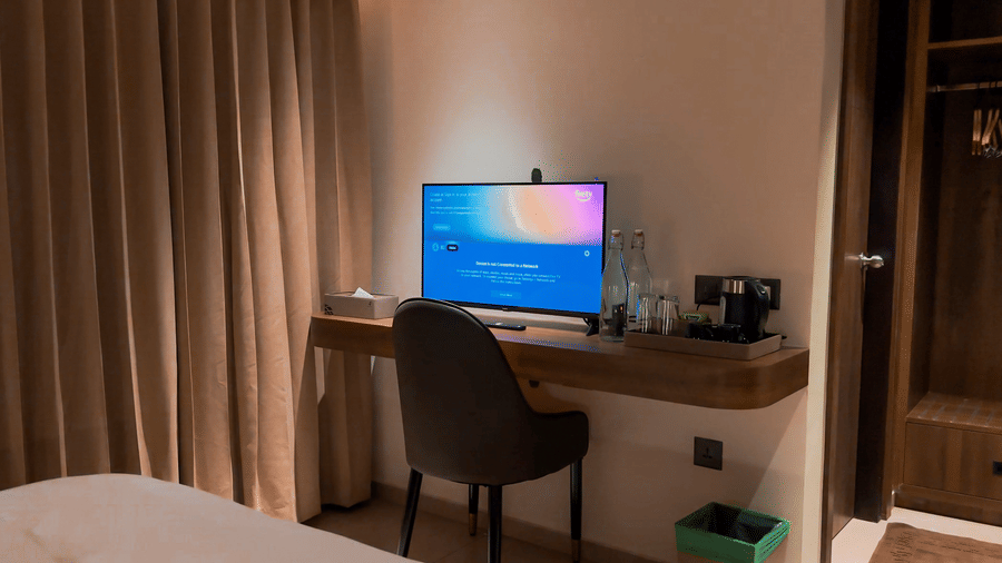 Close up of a chair and a desk with a smart TV kept on the table along with water bottles and refreshments in the Deluxe Room at F Hotels, Bengaluru.