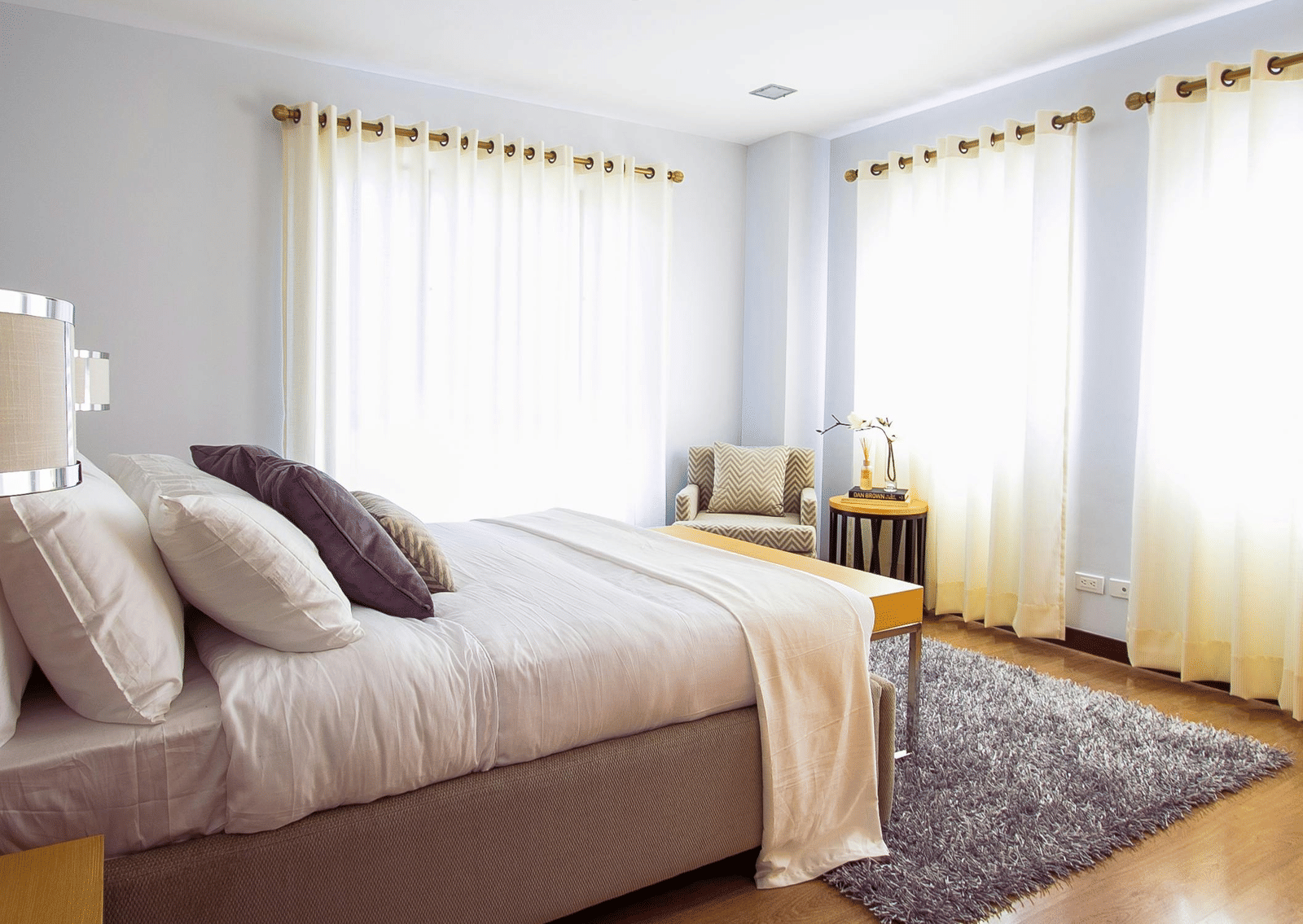 A neatly arranged bedroom with a large bed, pillows, side tables, armchair, and soft natural light filtering through curtains.