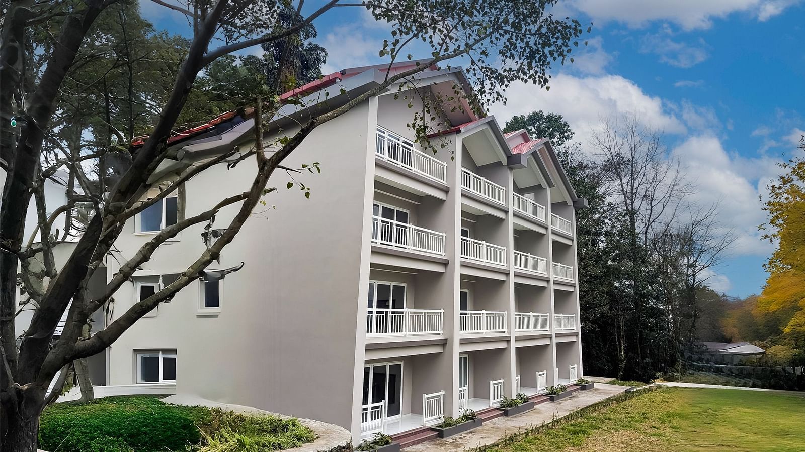 A multi-story building at Yashshree Resorts & Spa, Kalimpong, with a white facade and numerous windows, set against a backdrop of lush green trees and a blue sky.