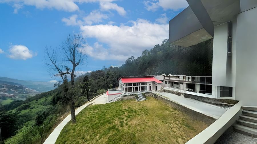 The hilltop view of Yashshree Resorts and Spa in Kalimpong featuring modern buildings with red roofs, terraced lawns, and panoramic mountain scenery under a partly cloudy sky.