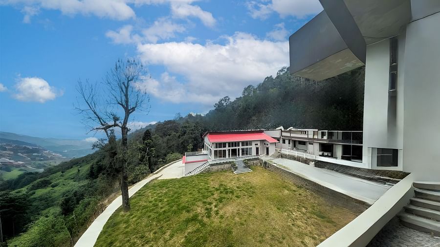 A panoramic view of Yashshree Resorts & Spa, Kalimpong, showing multiple buildings built into the hillside, overlooking a vast valley and distant mountains under a blue sky.