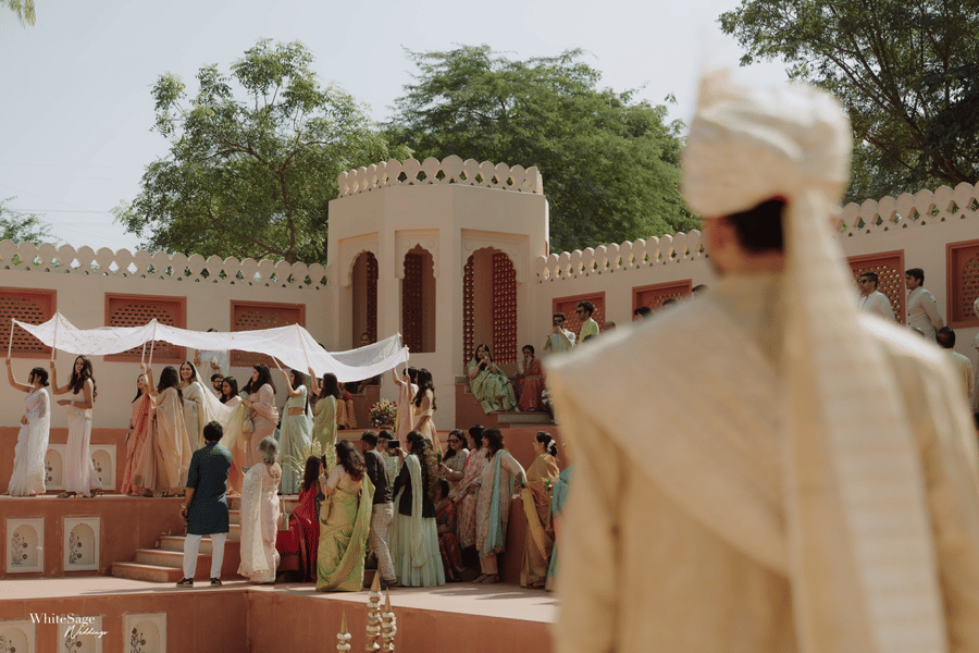 Groom wearing a beige turban watching the bride arrive under a white canopy at Umaid Palace.