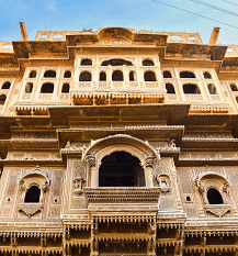 Ornate sandstone building facade with intricate carvings, balconies, and arched windows, viewed from below against a sky.