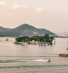 Lake Pichola view with Jag Mandir palace on island, boat in water, hills in background.