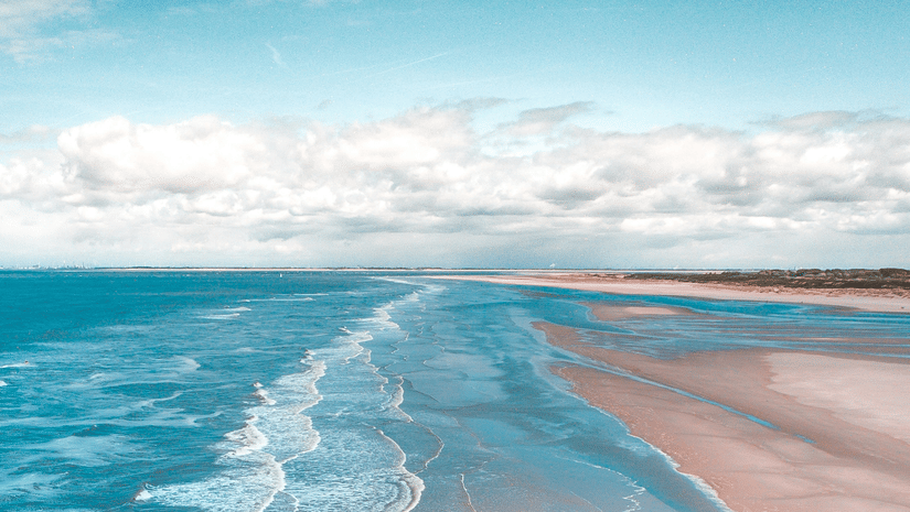 An aerial view of a sandy beach with gentle waves rolling onto the shore, under a bright blue sky with scattered clouds.