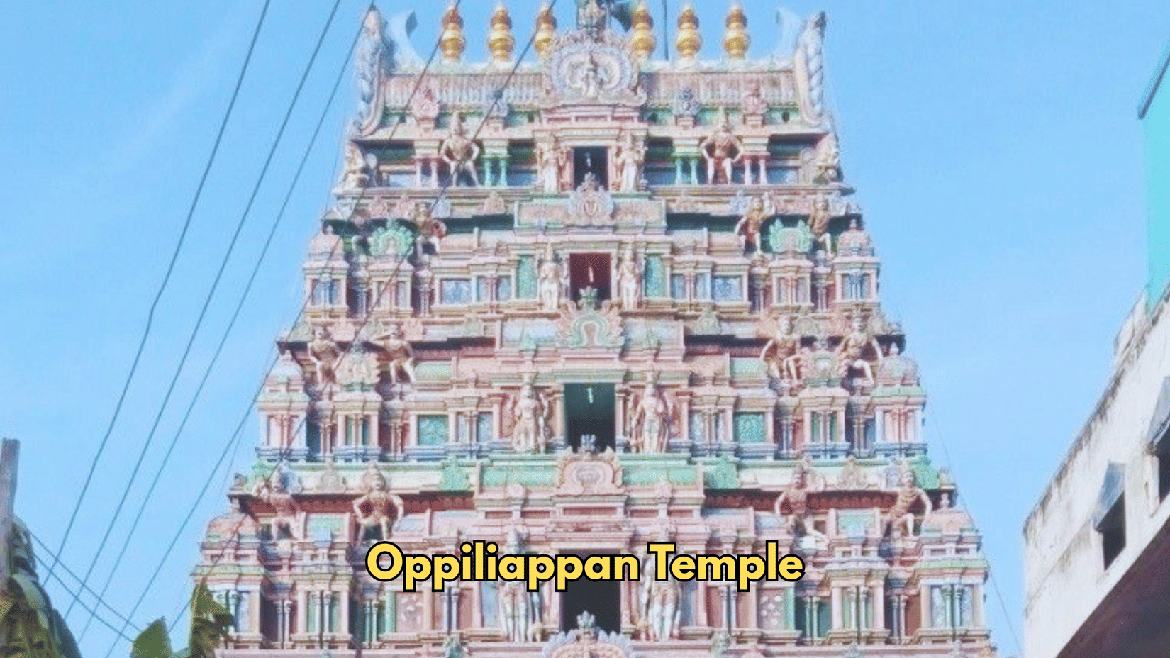 The entrance tower of Oppiliappan Temple features rows of carvings. A wall stands to the side of the structure.
