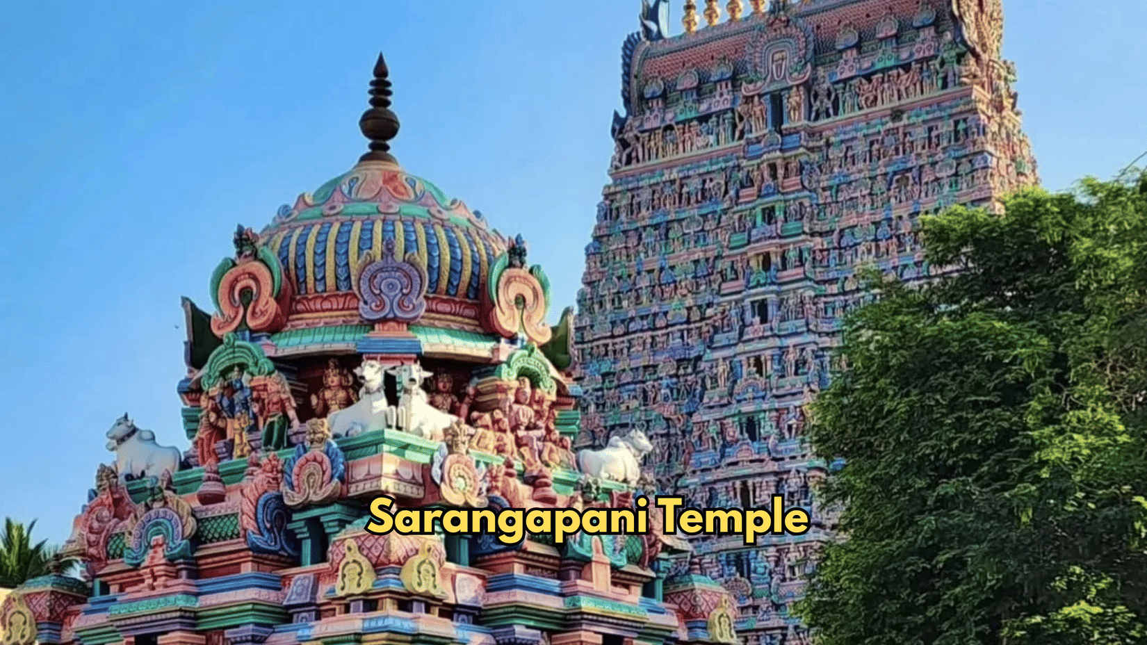 A dome stands beside a tower at Sarangapani Temple. Sculptures cover the tiers of the architecture. Foliage grows next to the structure.