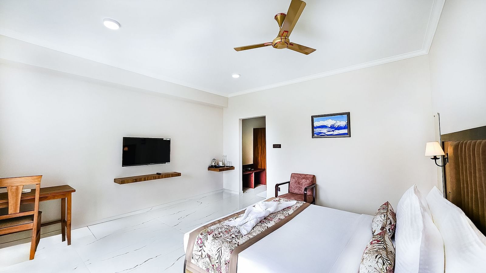 A simple hotel room with white walls, a large bed, a TV mounted on the wall, a wooden desk and chair, and a ceiling fan.