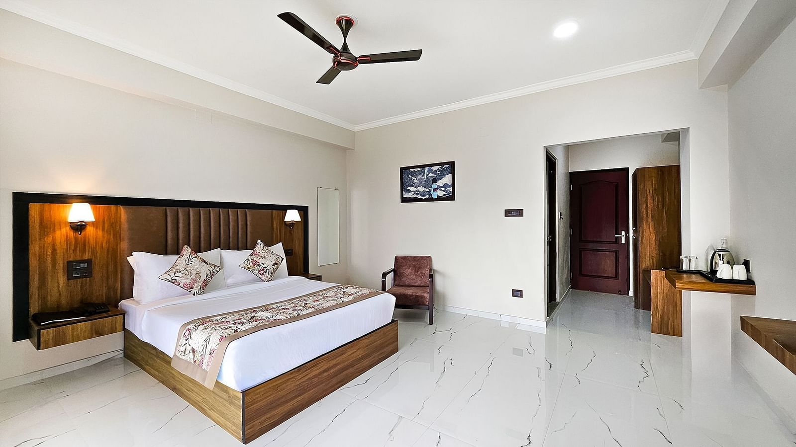  A spacious bedroom at Yashshree Resort & Spa Kalimpong features a large bed with a wooden headboard, a ceiling fan, a single armchair, and a glimpse of a hallway.
