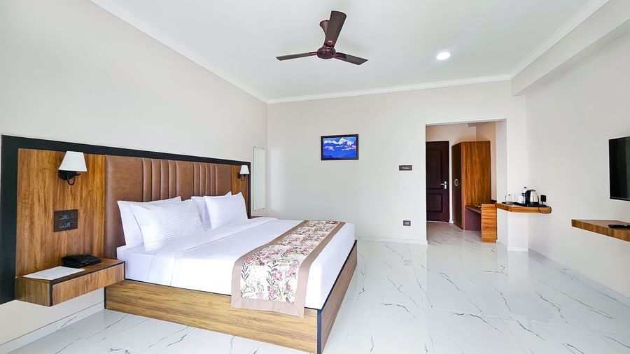 A premium executive room at Yashshree Resorts and Spa in Kalimpong featuring a spacious layout with a queen-size bed, a desk, a flat screen television, an almirah, a ceiling fan, and a painting on the wall.