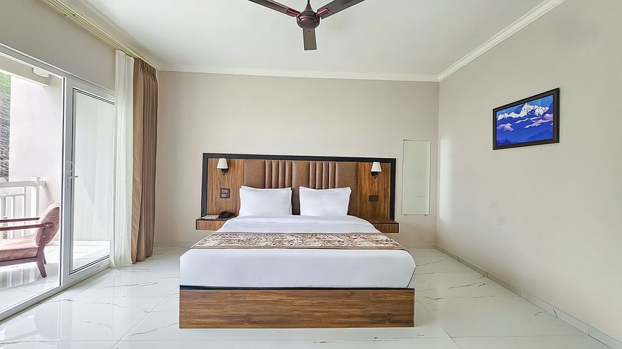 A bright bedroom at Yashshree Resort & Spa Kalimpong features a large bed with a wooden headboard, a ceiling fan, and a balcony to the left.