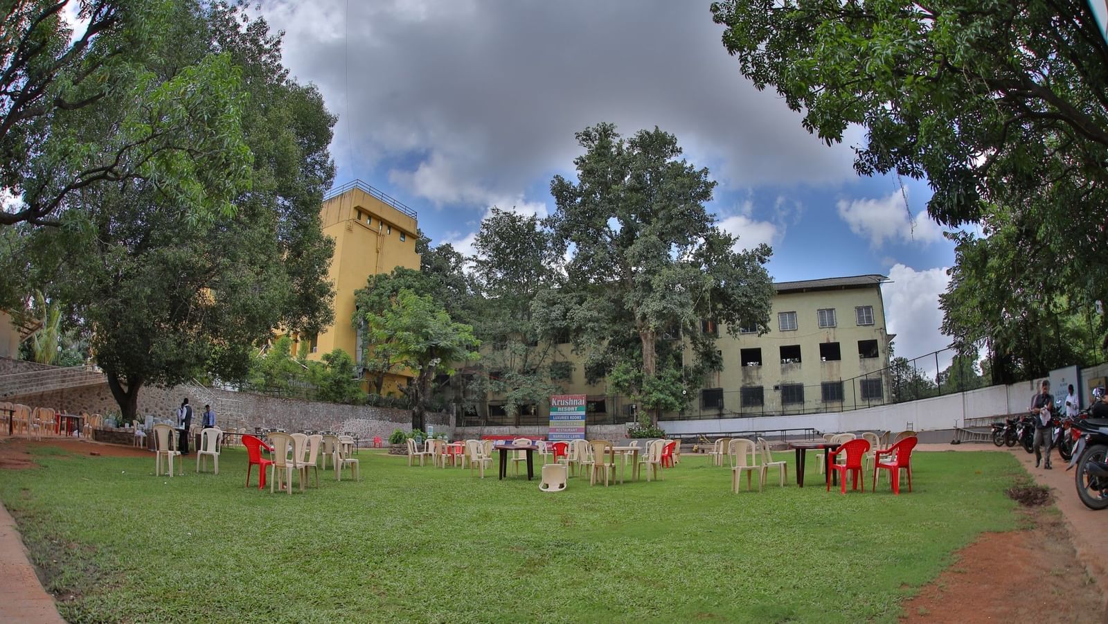 Green lawn at Krushnai Resort with scattered chairs and tables under a cloudy sky, surrounded by tall trees and distant buildings.