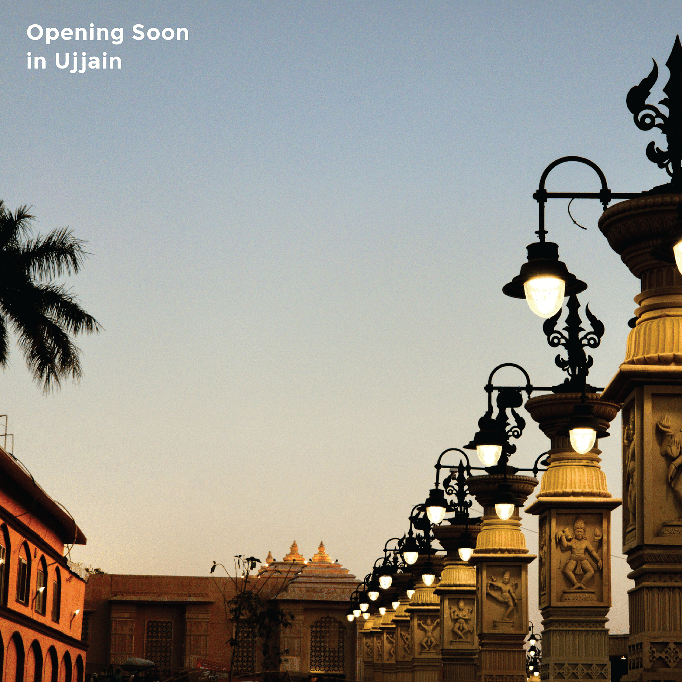 Ornate, illuminated street lamps line a walkway at dusk with traditional Indian architecture visible in the background and text - Opening Soon in Ujjain.