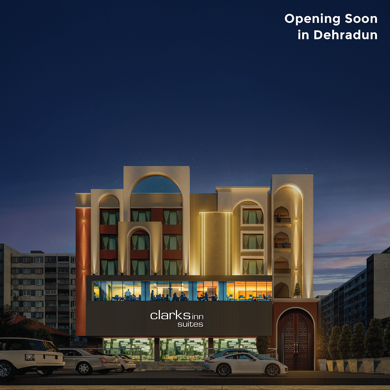 The illuminated facade of a modern Clarks Inn Suites is shown at dusk, with vehicles parked outside and text - Opening Soon in Dehradun.