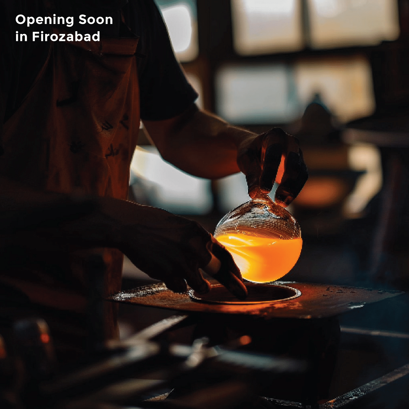 A persons hands are shaping molten orange glass over a heat source in a dimly lit workshop with text - Opening Soon in Firozabad.