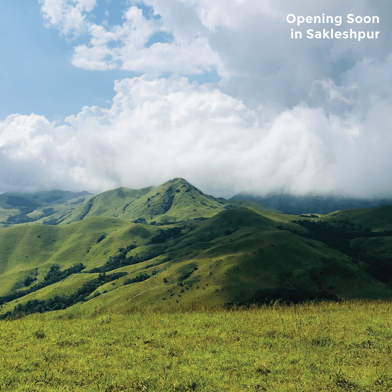 A scenic view of rolling green hills and valleys under a partly cloudy sky, with the text Opening Soon in Sakleshpur at the top right.