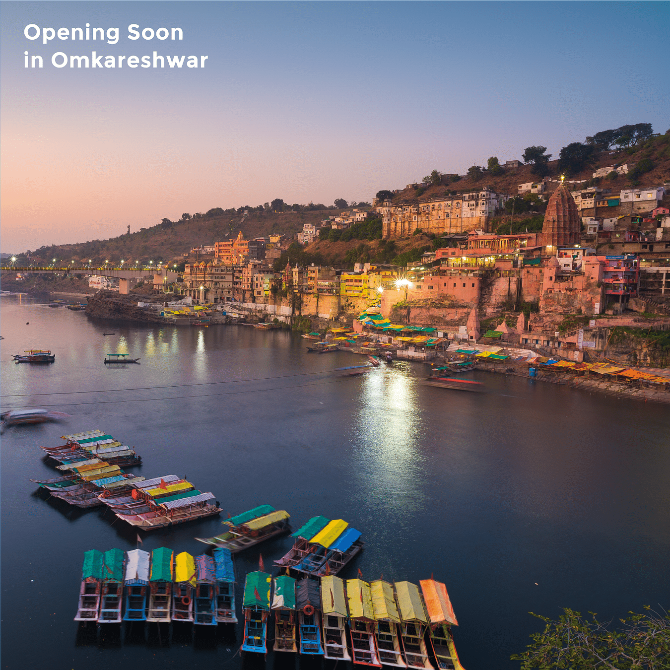 An aerial view of the lake in the evening with boats on the bank and temple on the other side of the bank as the text on the image says 'Opening Soon in Omkareshwar'