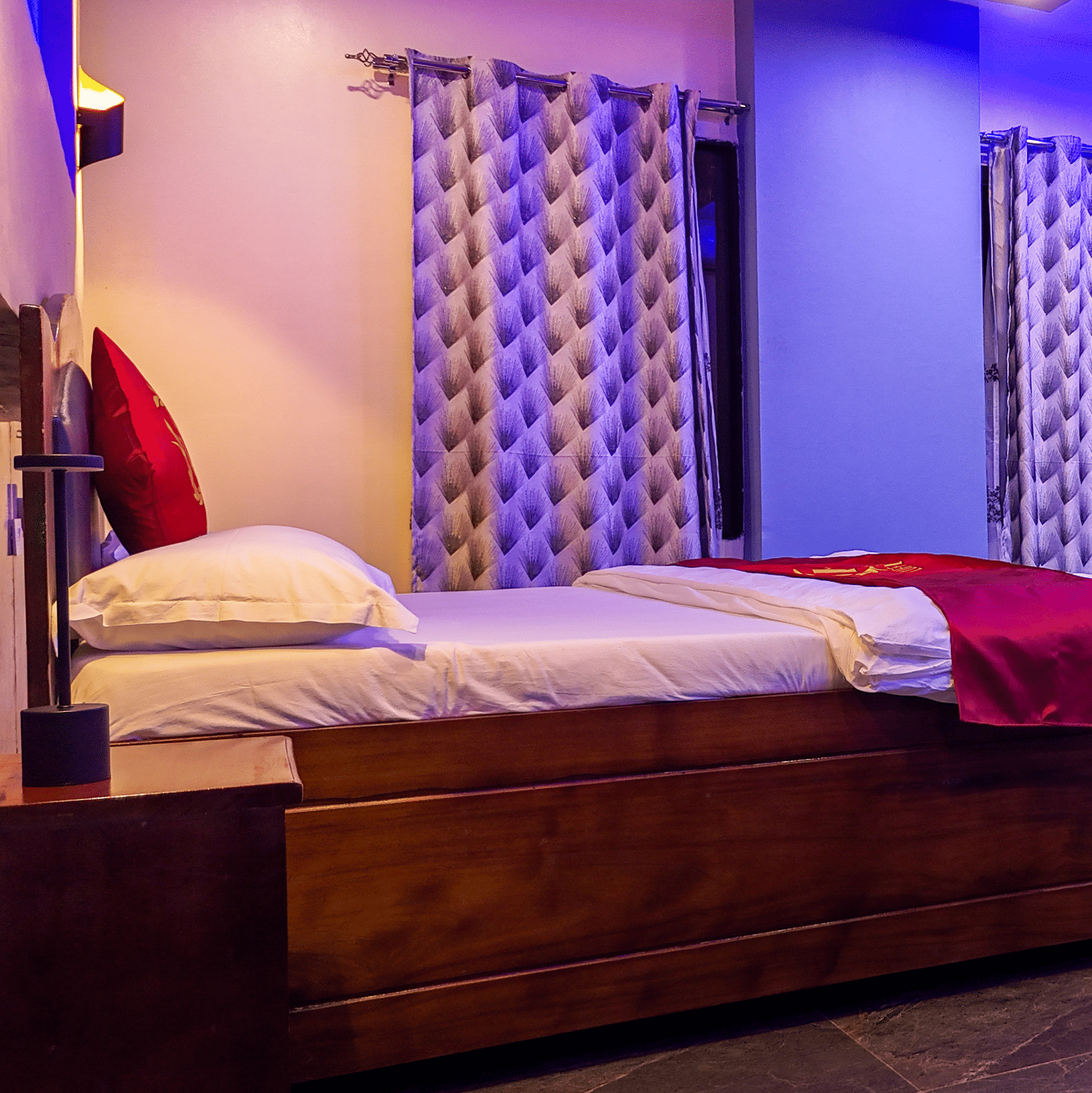 Interior view of a spacious room with a bed, small table and 2 chairs, patterned curtains, and vibrant purple lighting - Luffu Club