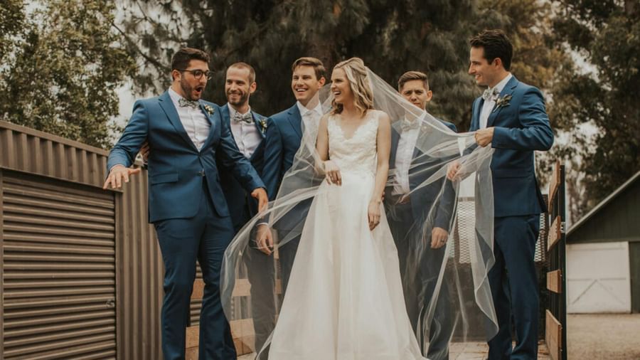 A wedding party, including the bride and groom and several attendants, stands together outdoors, smiling