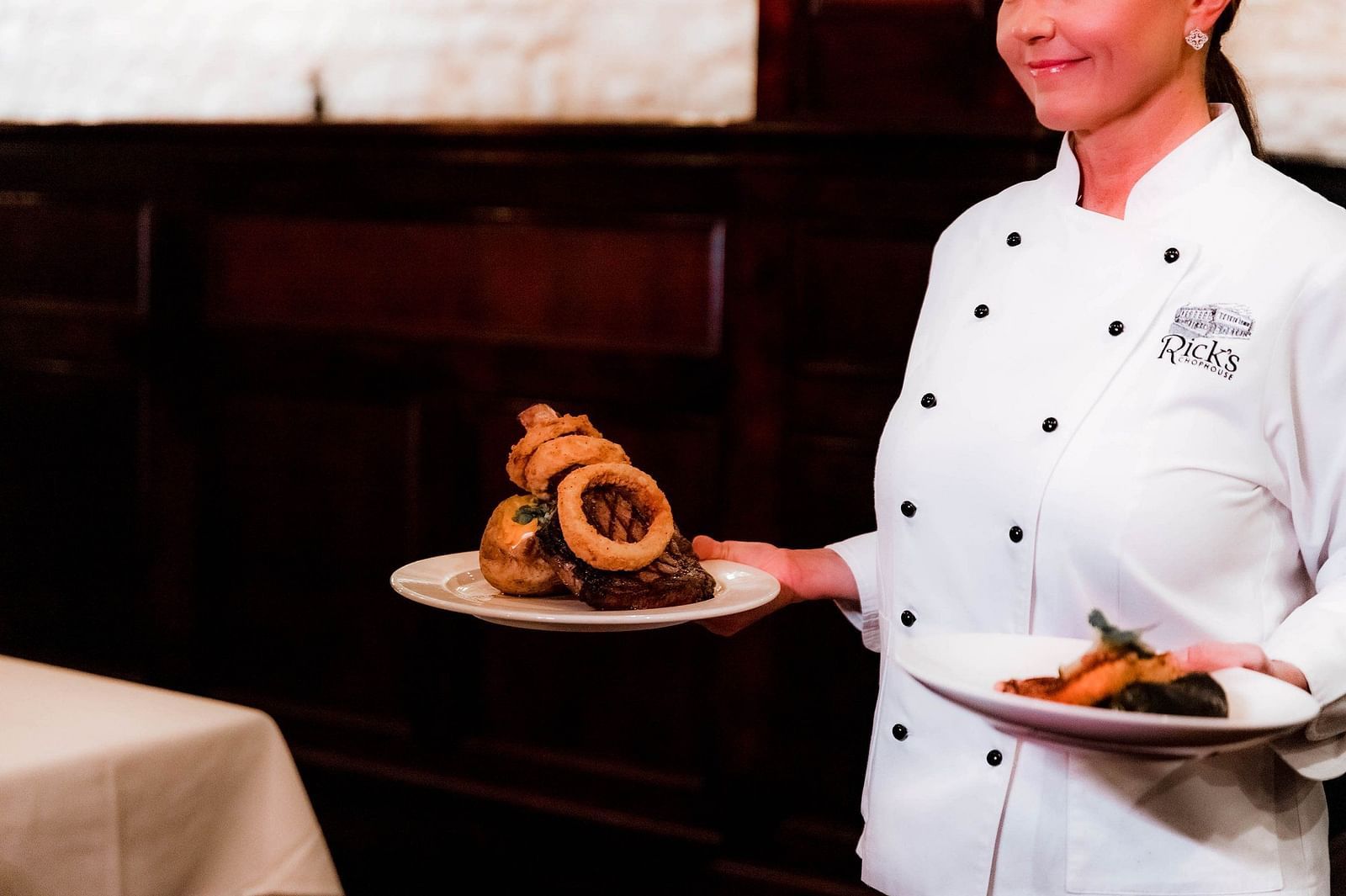 Chef standing carrying two plates in two hands with dishes on them wearing a chef coat  and table corner can be seen in the background