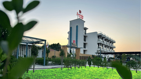 A modern hotel building is seen beyond a green lawn, with plants in the foreground and a clear sky above - Lords SKD Resort, Salangpur