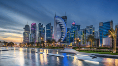 A cityscape at dusk with modern high-rise buildings illuminated along a waterfront. The buildings reflect in the water, and palm trees are visible along the shore.