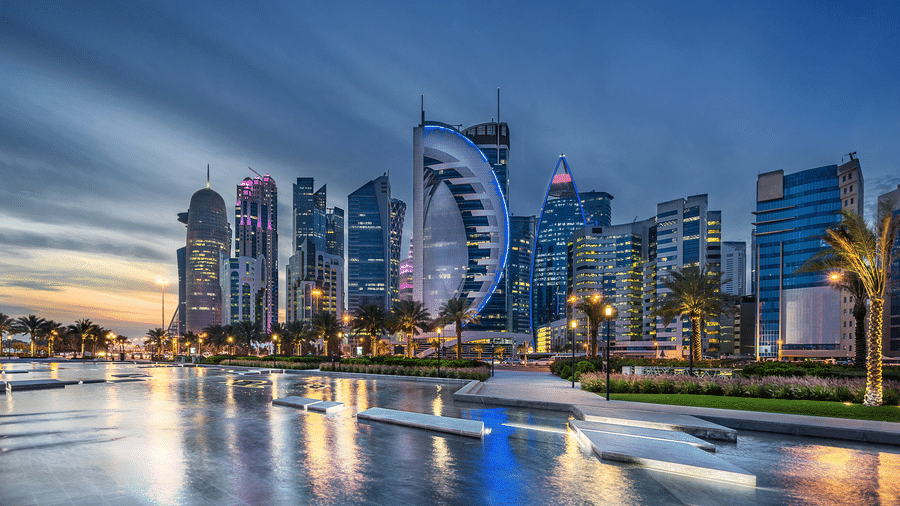 A cityscape at dusk with modern high-rise buildings illuminated along a waterfront. The buildings reflect in the water, and palm trees are visible along the shore - The Corniche in Doha, Qatar