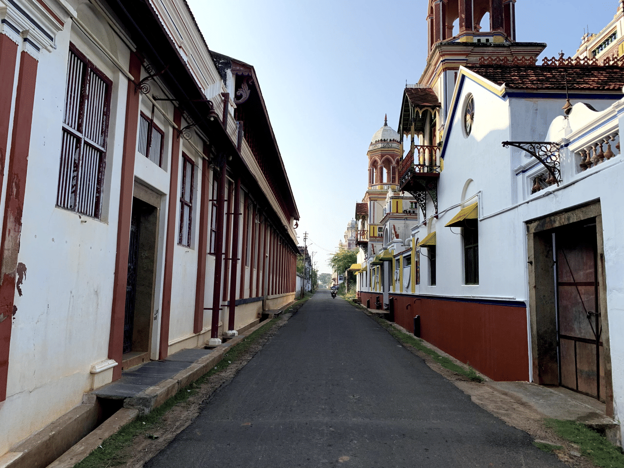 A narrow, paved street stretches between traditional white and red buildings with wooden frames and small balconies under a clear sky.