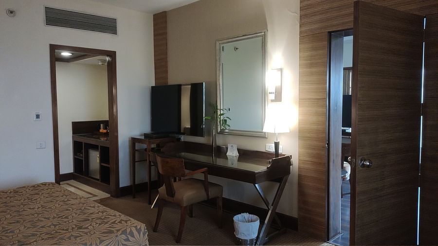 A neutral-toned hotel room featuring a desk with a mirror and TV, a chair, and an open door to an adjacent space - The Retreat Hotel and Convention Centre