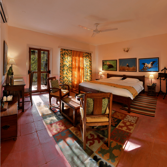 Bright heritage bedroom with wooden bed seating area patterned floor rugs artwork and long windows allowing natural light throughout the room at Neemrana’s Glasshouse on The Ganges offering a welcoming and restful environment