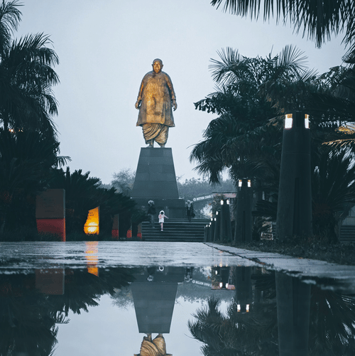 A golden statue is reflected in the still waters of a peaceful park at dusk.