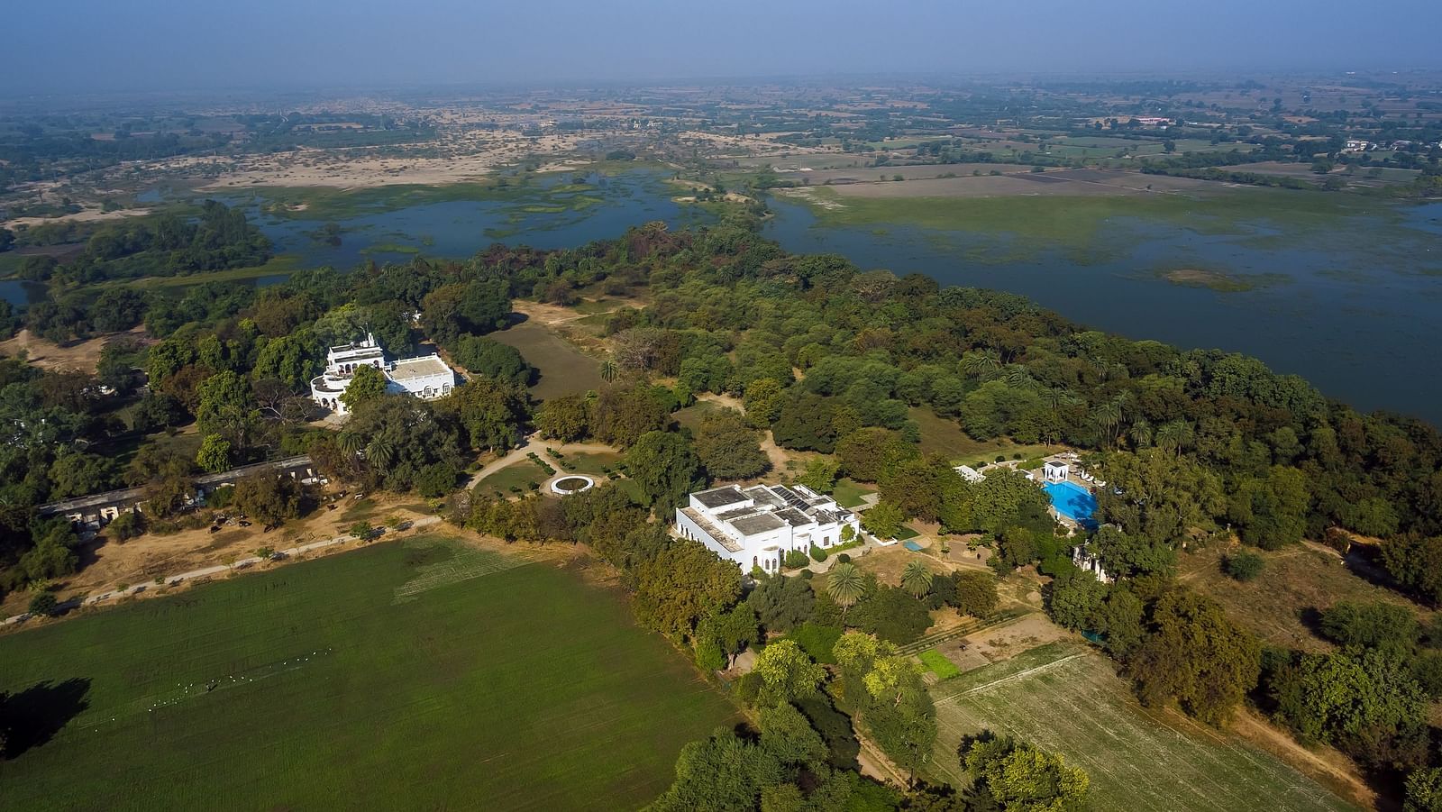 An aerial view of two white buildings nestled among lush greenery and a nearby lake- Shahpura Bagh.