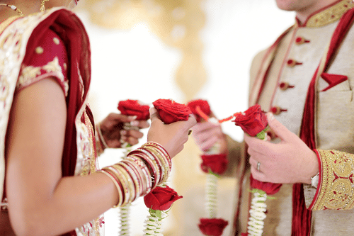 A close-up of a bride and groom's hands, with the bride's hand decorated with henna, holding red roses together.