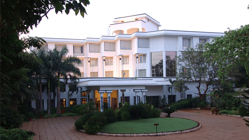 Facade view of Sangam Hotel, Thanjavur, with a pathway, a manicured garden and trees in view - Thanjavur Hotel Contact