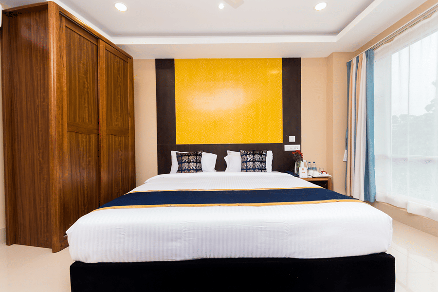 Suite featuring a plush bed with pillows, cushions, and a bed runner with a wardrobe nearby facing the window on the other side at Central Courtyard Boutique Hotel, Siliguri