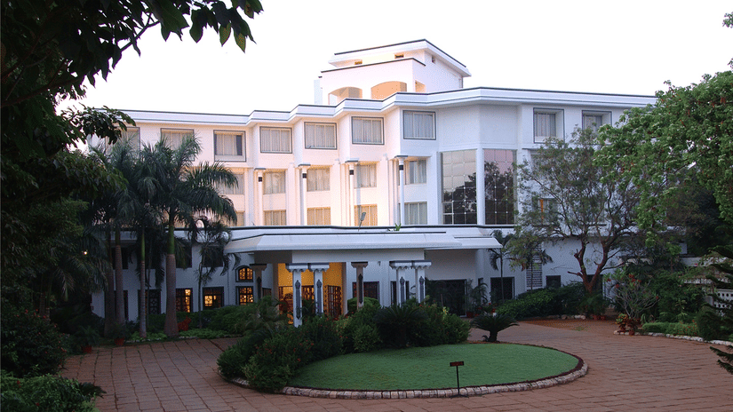 Facade view of Sangam Hotel, Thanjavur with a pathway, a manicured garden and trees in view.