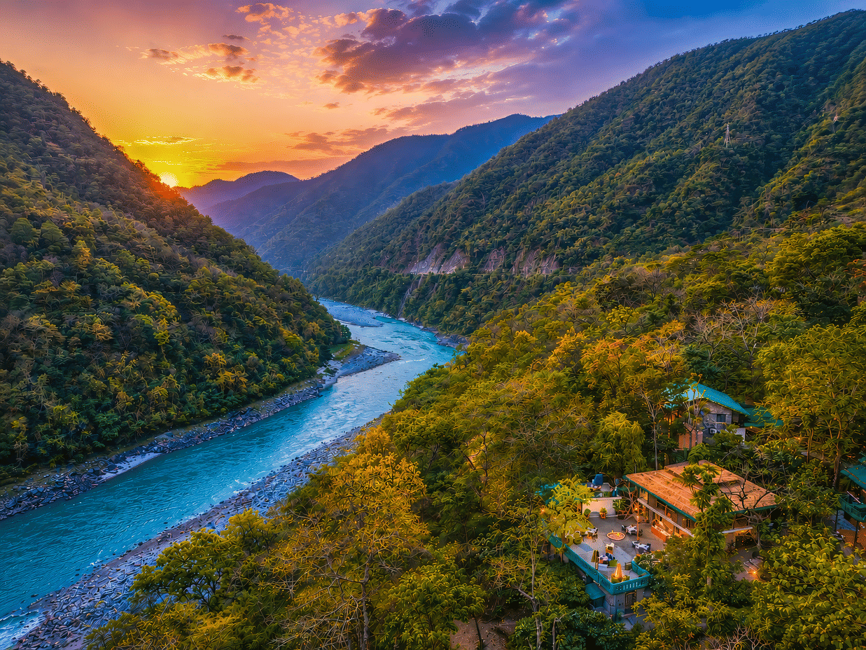 Aerial view of the Neemran's Glasshouse on The Ganges, Rishikesh, surrounded by lush greenery by the river banks, along with a mesmerising sunset in the background.