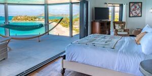 A bedroom with a large bed and a hammock, offering a panoramic ocean view through a large window.