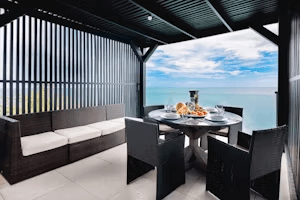 An outdoor dining area under a shaded pergola offers a view of the ocean in the distance.